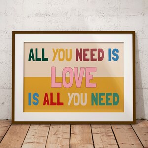 A framed Beatles print leans against a wooden floor. The music poster features a split yellow and beige background with the lyrics 'All You Need is Love' written is multiple colours.