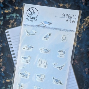 Peut inclure: Une feuille d'autocollants avec des illustrations de mouettes dans diverses poses. Les autocollants sont principalement bleus et blancs, sur un fond bleu clair. Le texte "Resting Cat" et "Seagull" est visible.
