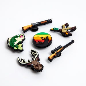 May include: A collection of hunting-themed shoe charms. The charms include a green and white fish, a brown and white deer head, a duck in flight, two rifles, and a round charm with a hunter silhouette.