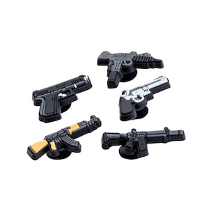 Op de afbeelding: Een verzameling van vijf zwart-grijze pistoolvormige bedels. De bedels tonen verschillende vuurwapenontwerpen, waaronder pistolen en geweren, sommige met zilveren of bruine accenten. Deze accessoires zijn waarschijnlijk voor decoratieve doeleinden.