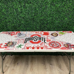 May include: A white folding table with a red and gray Ohio State University design. The table features the Ohio State logo, Buckeye mascot, and other graphics. It has a series of cup holders and is supported by metal legs.