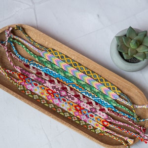 Peut inclure: Un plateau en bois contenant une sélection de bracelets d'amitié colorés. Les bracelets sont faits de fils de différentes couleurs et présentent des motifs complexes.