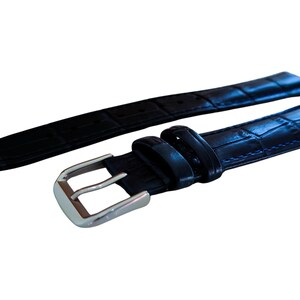 K&ouml;nnte beinhalten: Ein schwarzes Uhrenarmband mit einem strukturierten, krokodilartigen Muster. Das Armband verf&uuml;gt &uuml;ber eine silberfarbene Schnalle und ist zur Befestigung an einem Zifferblatt konzipiert. Das Armband besteht aus einem strapazierf&auml;higen Material.