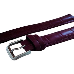 K&ouml;nnte beinhalten: Ein burgunderfarbenes Lederarmband mit einer silberfarbenen Schnalle. Das Armband hat ein strukturiertes, krokodilartiges Muster. Das Uhrenarmband wird vor einem wei&szlig;en Hintergrund gezeigt.