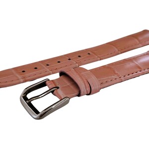 K&ouml;nnte beinhalten: Hellrosa Uhrenarmband mit Krokodilmuster. Das Armband hat eine silberfarbene Schnalle und mehrere L&ouml;cher zur Anpassung. Das Uhrenarmband besteht aus einem strukturierten Material.