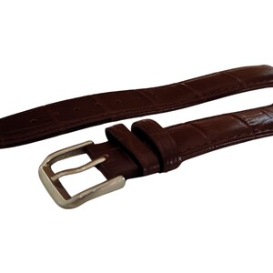 K&ouml;nnte beinhalten: Ein braunes Uhrenarmband mit einem strukturierten, krokodilartigen Muster. Das Armband ist in zwei Teile geteilt, mit einer silberfarbenen Schnalle an einem Ende. Das Leder hat eine satte, dunkelbraune Farbe.