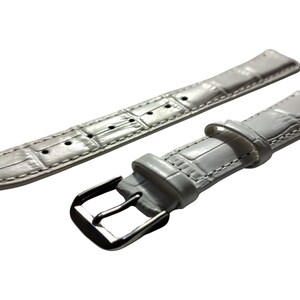 K&ouml;nnte beinhalten: Ein hellgraues Uhrenarmband mit einem strukturierten, krokodilartigen Muster. Das Armband besteht aus zwei Teilen, einem mit einer Schnalle und einem mit mehreren L&ouml;chern zur Anpassung. Die Metallschnalle ist silberfarben.