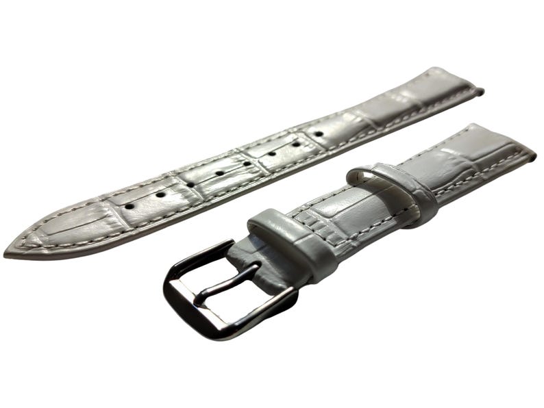 K&ouml;nnte beinhalten: Ein hellgraues Uhrenarmband mit einem strukturierten, krokodilartigen Muster. Das Armband besteht aus zwei Teilen, einem mit einer Schnalle und einem mit mehreren L&ouml;chern zur Anpassung. Die Metallschnalle ist silberfarben.