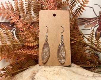 May include: A pair of silver dangle earrings with a clear, iridescent, oval-shaped resin pendant. The earrings are displayed on a brown card.