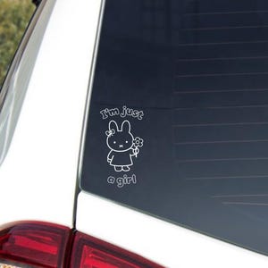 May include: White vinyl decal featuring a cartoon bunny holding a flower, with the text "I'm just a girl". The decal is applied to a car window. The bunny has a bow and is wearing a dress.