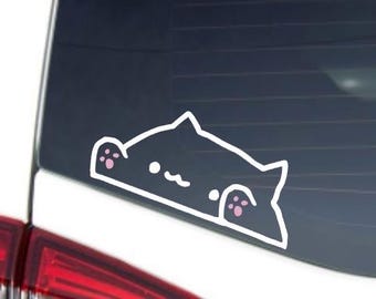 Puede incluir: Una pegatina de contorno blanco de un gato de dibujos animados con almohadillas rosas, asomándose por encima de una superficie. La pegatina está en una ventana de coche, con la luz trasera roja visible.
