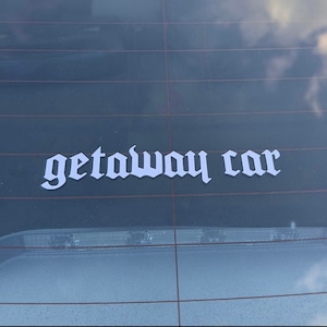 Può includere: Un adesivo in vinile bianco con le parole "getaway car" in un font gotico su un finestrino di un'auto.
