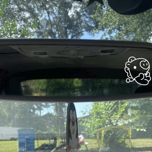 Op de afbeelding: Een witte achteruitkijkspiegel van een auto met een witte cartoon-varkenssticker op het glas. De spiegel reflecteert een groene boom en een blauwe lucht.