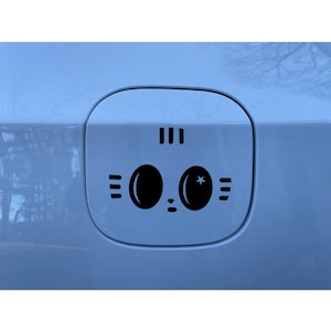 Puede incluir: Una calcoman&iacute;a de vinilo negro aplicada en la tapa del combustible de un veh&iacute;culo azul claro. La calcoman&iacute;a presenta un rostro estilizado con dos ojos ovalados grandes, uno con una peque&ntilde;a estrella, tres l&iacute;neas verticales arriba.