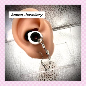 May include: A close-up of a white and black ear gauge with a silver chain attached. The chain has small silver hearts hanging from it. The ear gauge is in a person's ear. The text "Acton Jewellery" is visible in the image.