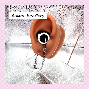 May include: A silver chain earring with a star charm hanging from the bottom. The earring is attached to a white ear cuff. The ear cuff is worn on a model ear.