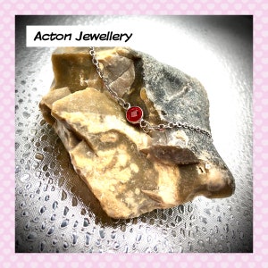 May include: A silver chain necklace with a small, round, red gemstone pendant. The necklace is displayed on a rough, brown rock.