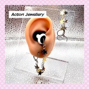 May include: A pair of silver star dangle earrings with gold star accents. The earrings are hanging from a model ear.