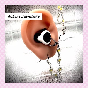 May include: A pair of silver earrings with a chain of small white flowers with yellow centres. The earrings are hanging from a person's ear. The earrings are a delicate and feminine accessory.