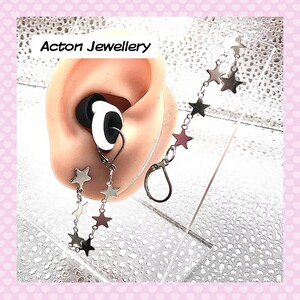 May include: A close-up of a silver star charm earring with a chain of stars hanging from it. The earring is attached to a person's earlobe. The image is captioned "Acton Jewellery".