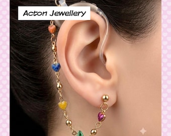 May include: A gold-toned hearing aid accessory chain featuring small, colorful enamel heart charms and round beads. The chain connects to a clear plastic hearing aid device. The text Acton Jewellery is visible in the upper left corner of the image.