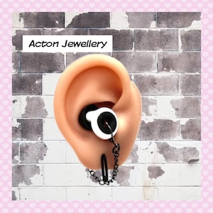 May include: A black chain earring with a white and black earbud-shaped charm. The earring is worn in a model ear.