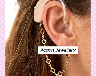 May include: A gold-toned metal chain accessory for a hearing aid, featuring a series of small, open-work clover-shaped links. The chain attaches to the beige hearing aid device worn behind the ear. The text Acton Jewellery is visible.