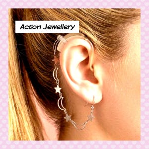 May include: A silver ear cuff featuring stars and crescent moons is attached to an ear with a hearing aid. The ear cuff has a chain that connects to a dangling earring with stars. The text "Acton Jewellery" is in a white box in the upper left corner.
