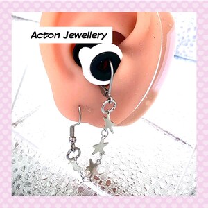 May include: A close-up of a silver star-shaped earring with a small ball at the end. The earring is attached to a clear plastic earlobe. The text "Acton Jewellery" is visible in the background.