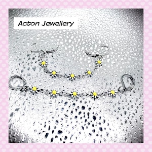 May include: A pair of silver chain earrings with yellow daisy charms. The earrings are on a white background with a water droplet pattern. The text "Acton Jewellery" is at the top of the image.