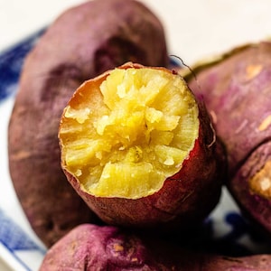 Op de afbeelding: Close-up van een gebakken zoete aardappel die open is gesneden, waardoor een heldergele, zachte binnenkant zichtbaar wordt. De aardappel wordt omringd door andere hele zoete aardappelen, allemaal met een diepe paarsrode schil.