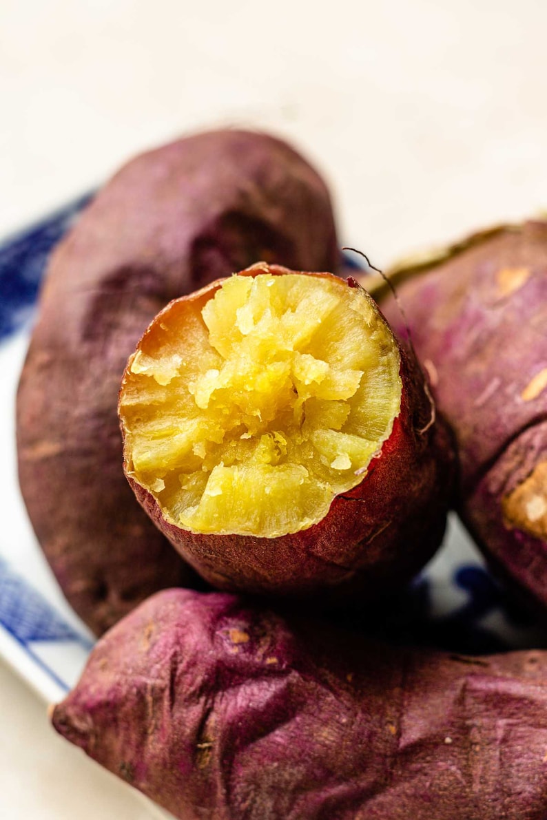 Op de afbeelding: Close-up van een gebakken zoete aardappel die open is gesneden, waardoor een heldergele, zachte binnenkant zichtbaar wordt. De aardappel wordt omringd door andere hele zoete aardappelen, allemaal met een diepe paarsrode schil.