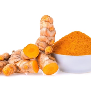 May include: A bowl of turmeric powder next to a group of turmeric root pieces, some of which are sliced in half, on a white background.