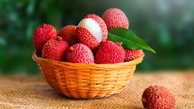 May include: A wicker basket filled with fresh lychees, some with the skin removed, revealing the white flesh. The basket is on a hessian surface.