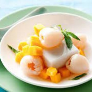 May include: A white bowl containing a white, creamy dessert topped with diced mango and lychees. The dessert is garnished with a green leaf.