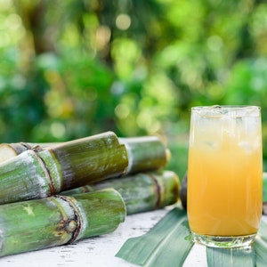 Peut inclure: Un verre de jus jaune avec des gla&ccedil;ons, &agrave; c&ocirc;t&eacute; de plusieurs tiges de canne &agrave; sucre vertes. L'image est prise sur un fond vert flou, sugg&eacute;rant un cadre naturel. La boisson est dans un verre transparent.