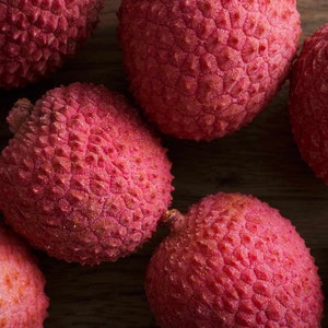 May include: Close-up of several red lychees with a textured, bumpy skin. The lychees are arranged on a wooden surface.