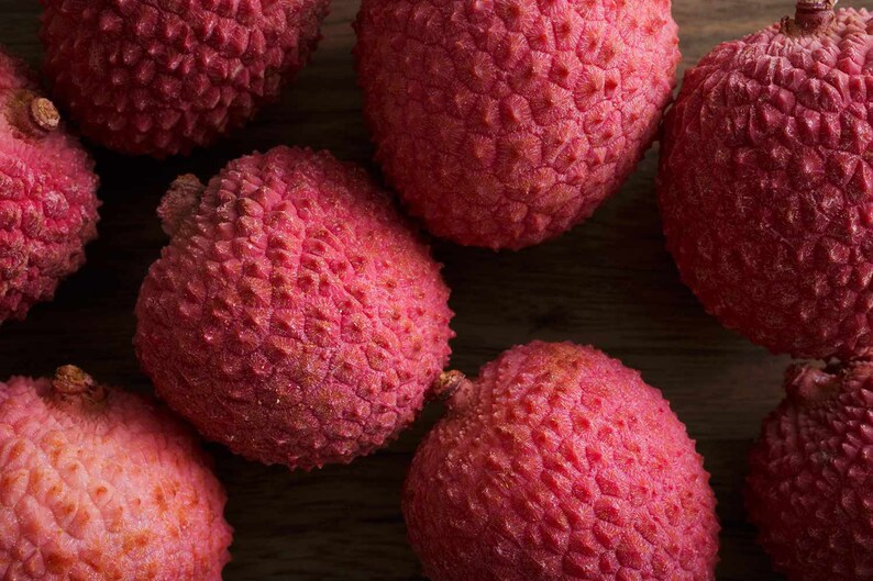 May include: Close-up of several red lychees with a textured, bumpy skin. The lychees are arranged on a wooden surface.