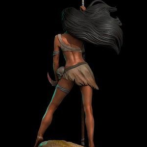 Puede incluir: Una mujer con el pelo largo y negro se para en una roca sosteniendo una lanza con una punta de plumas blancas. Lleva una falda de cuero marr&oacute;n y una correa de cuero alrededor de su muslo izquierdo.