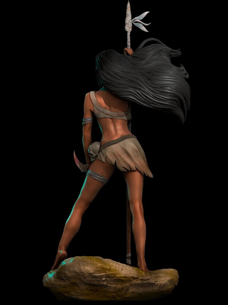 Puede incluir: Una mujer con el pelo largo y negro se para en una roca sosteniendo una lanza con una punta de plumas blancas. Lleva una falda de cuero marr&oacute;n y una correa de cuero alrededor de su muslo izquierdo.