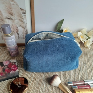 Può includere: Un beauty case blu in spugna con cerniera bianca, aperto per rivelare l'interno. Un pennello per il trucco, rossetto e una palette per il trucco sono accanto alla borsa. Una bottiglia di profumo e un quadro incorniciato sono sullo sfondo.
