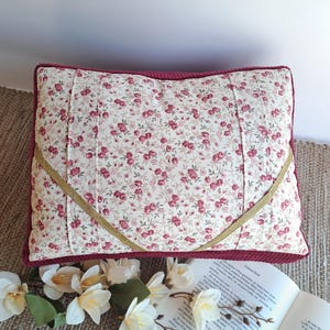 Peut inclure: Coussin rectangulaire à motifs floraux avec de petites fleurs rouges sur fond blanc. Bordure en velours côtelé bordeaux et détail ruban doré. Le coussin est posé sur un tapis tissé.