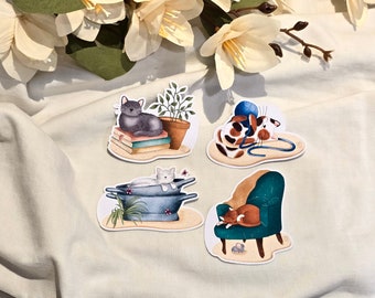 Op de afbeelding: Vier stickers met katten als thema. Eén toont een grijze kat op boeken met een plant in een pot. Een andere toont een lapjeskat met garen. Een derde toont een witte kat in een pot. De laatste toont een oranje kat op een tealkleurige fauteuil.