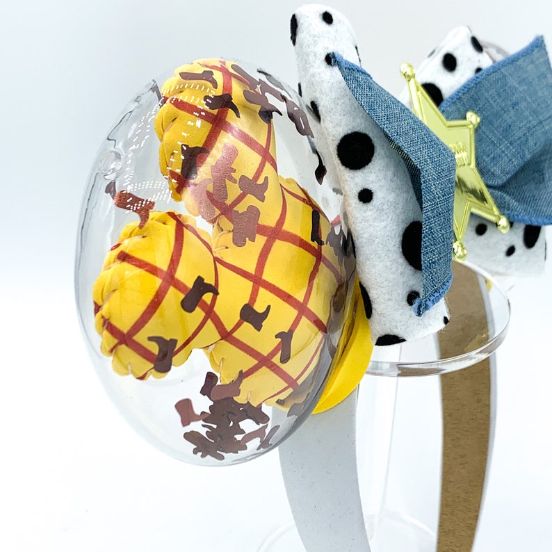 May include: A clear, oval-shaped ornament featuring a yellow figure with a red plaid pattern and brown accents. A white headband with a dalmatian-spotted bow and a gold star-shaped badge completes the design. The ornament is on a clear stand.