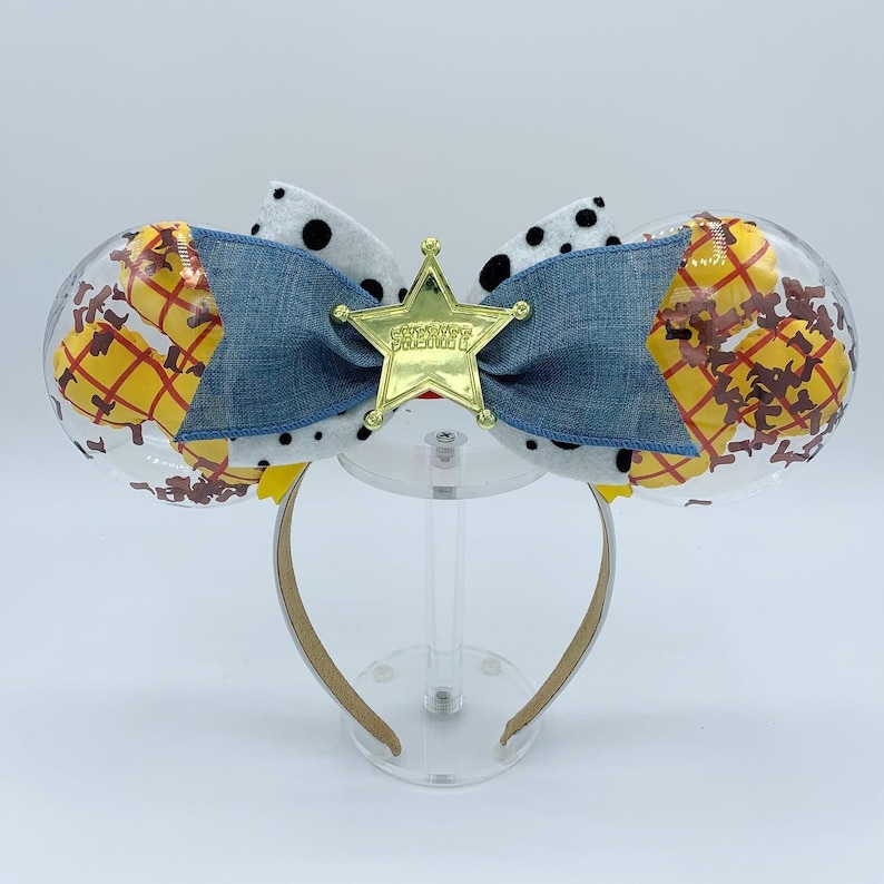 May include: A themed headband featuring clear, round ears filled with a Woody-inspired design. A denim bow is centered with a gold star badge that reads "SHERIFF WOODY". The headband has a tan band.