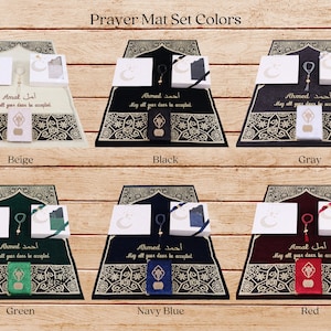 May include: A set of prayer mats in six different colors: beige, black, gray, green, navy blue, and red. Each mat features a traditional Islamic design with gold accents and includes a matching prayer rug, prayer beads, and a small box. The text on the mats reads "Ahmed" and "Amal" in Arabic, with the English translation "May all your deeds be accepted."