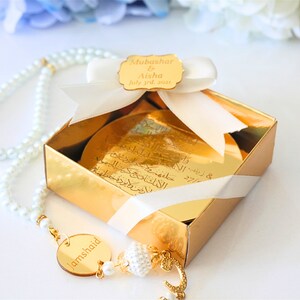 May include: A gold gift box with a white ribbon and a gold tag that says "Mubashar & Aisha July 3rd, 2021". A white pearl necklace with a gold pendant that says "Jamshaid" is in front of the box.