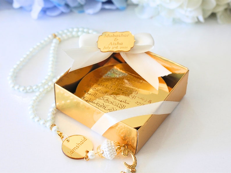 May include: A gold gift box with a white ribbon and a gold tag that says "Mubashar & Aisha July 3rd, 2021". A white pearl necklace with a gold pendant that says "Jamshaid" is in front of the box.