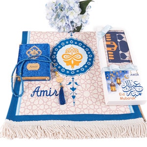 May include: An Islamic prayer set featuring a blue velvet Quran, prayer beads, and a prayer rug with a geometric pattern. The set includes a gift box with "Eid Mubarak" text and a tassel. A blue floral arrangement is in the background.