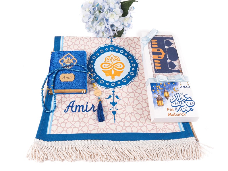 May include: An Islamic prayer set featuring a blue velvet Quran, prayer beads, and a prayer rug with a geometric pattern. The set includes a gift box with "Eid Mubarak" text and a tassel. A blue floral arrangement is in the background.